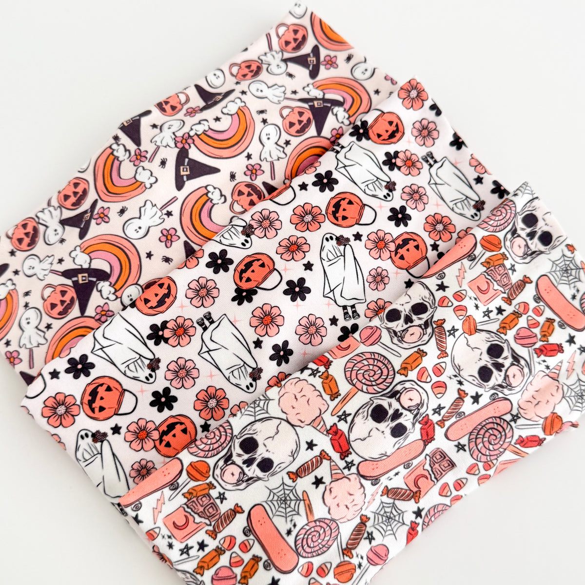 Three folded fabric swatches of the Halloween Knotted Tie Headband prints — pastel witch hat and rainbow print, ghost and pumpkin floral print, and skull and candy print