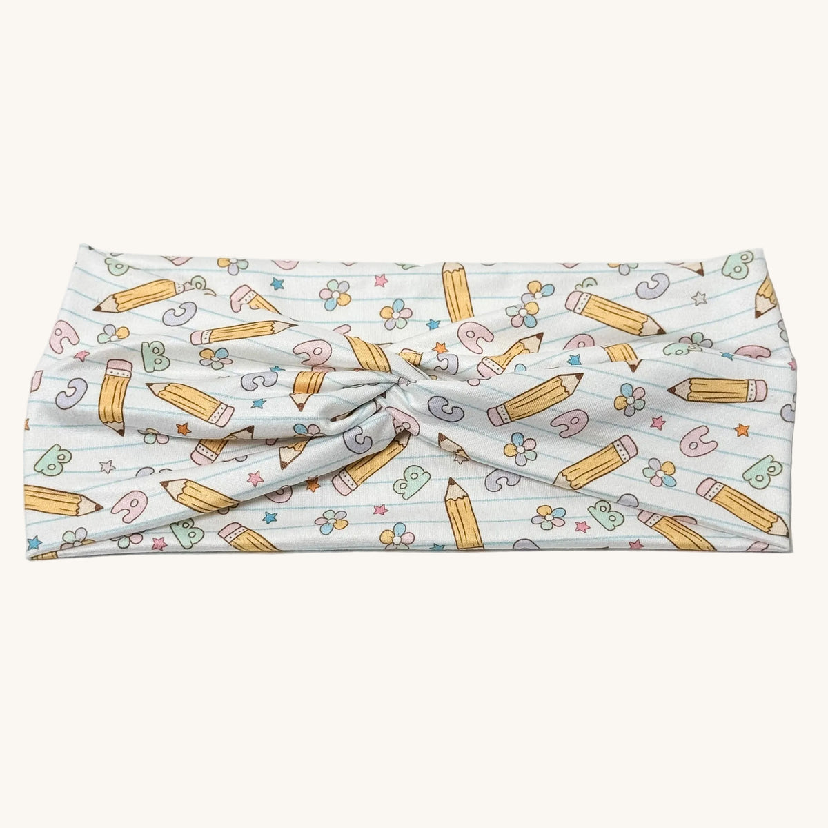 A knotted fabric headband featuring a playful school-themed pattern with yellow pencils, pastel flowers, and numbers on a white background.