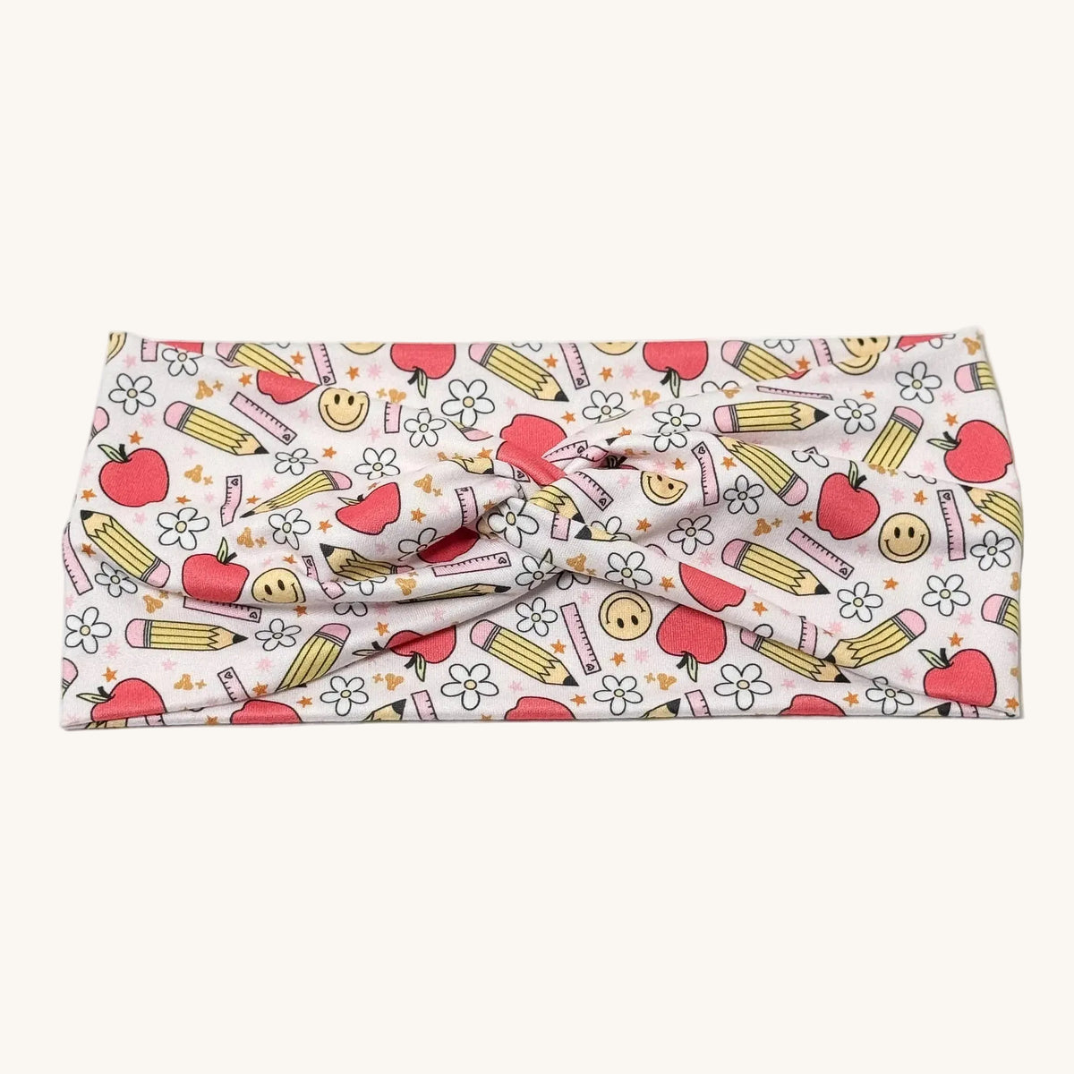 A fabric headband with a school-themed pattern featuring red apples, yellow pencils, smiley faces, and small flowers on a white background.