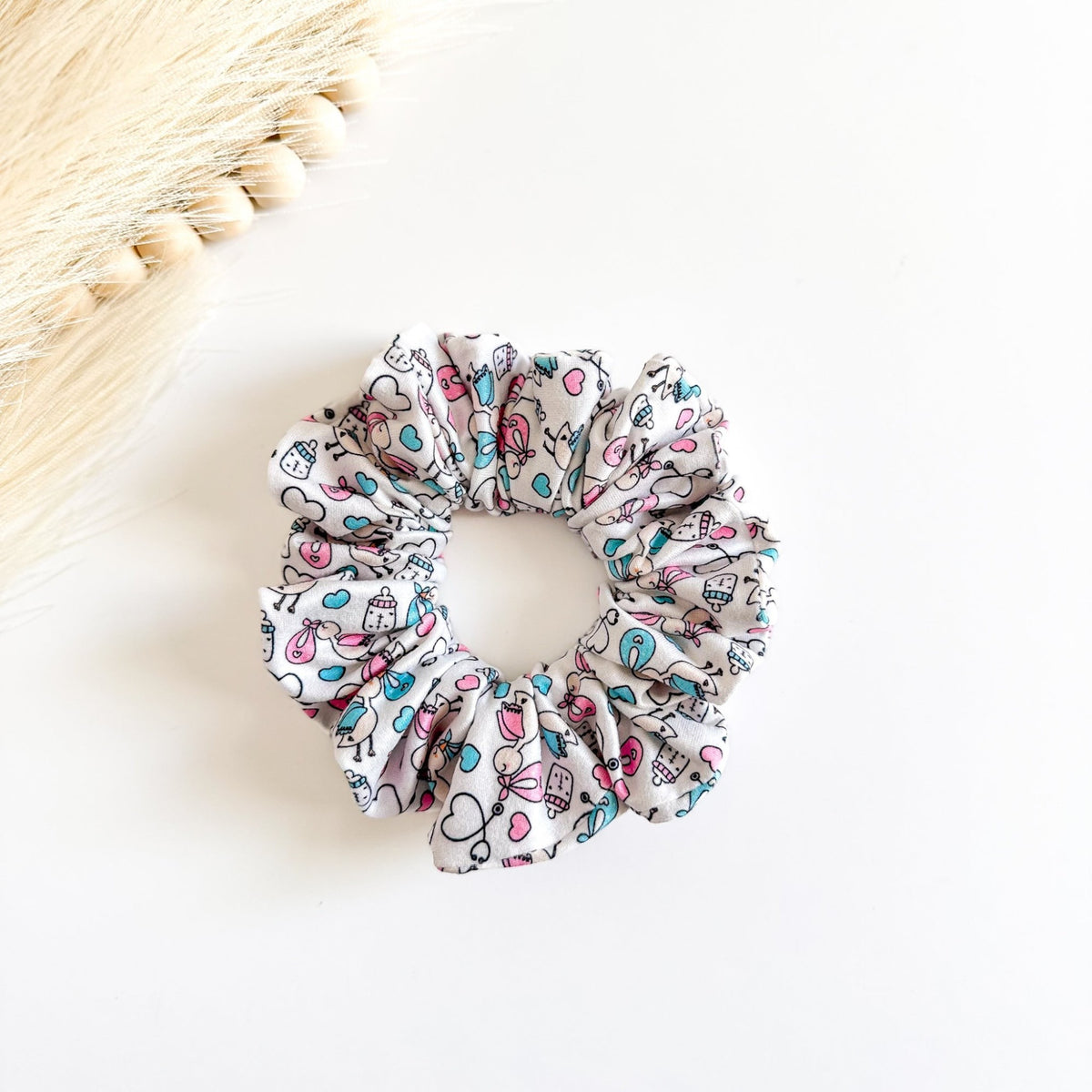 Headband Studio white scrunchie with a playful medical print featuring baby bottles, hearts, and birds in pink and blue.