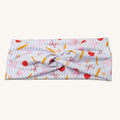 A knotted fabric headband featuring a pastel blue and white striped background with a playful pattern of red apples, yellow pencils, pink bows, and orange stars, displayed on a clean off-white background.
