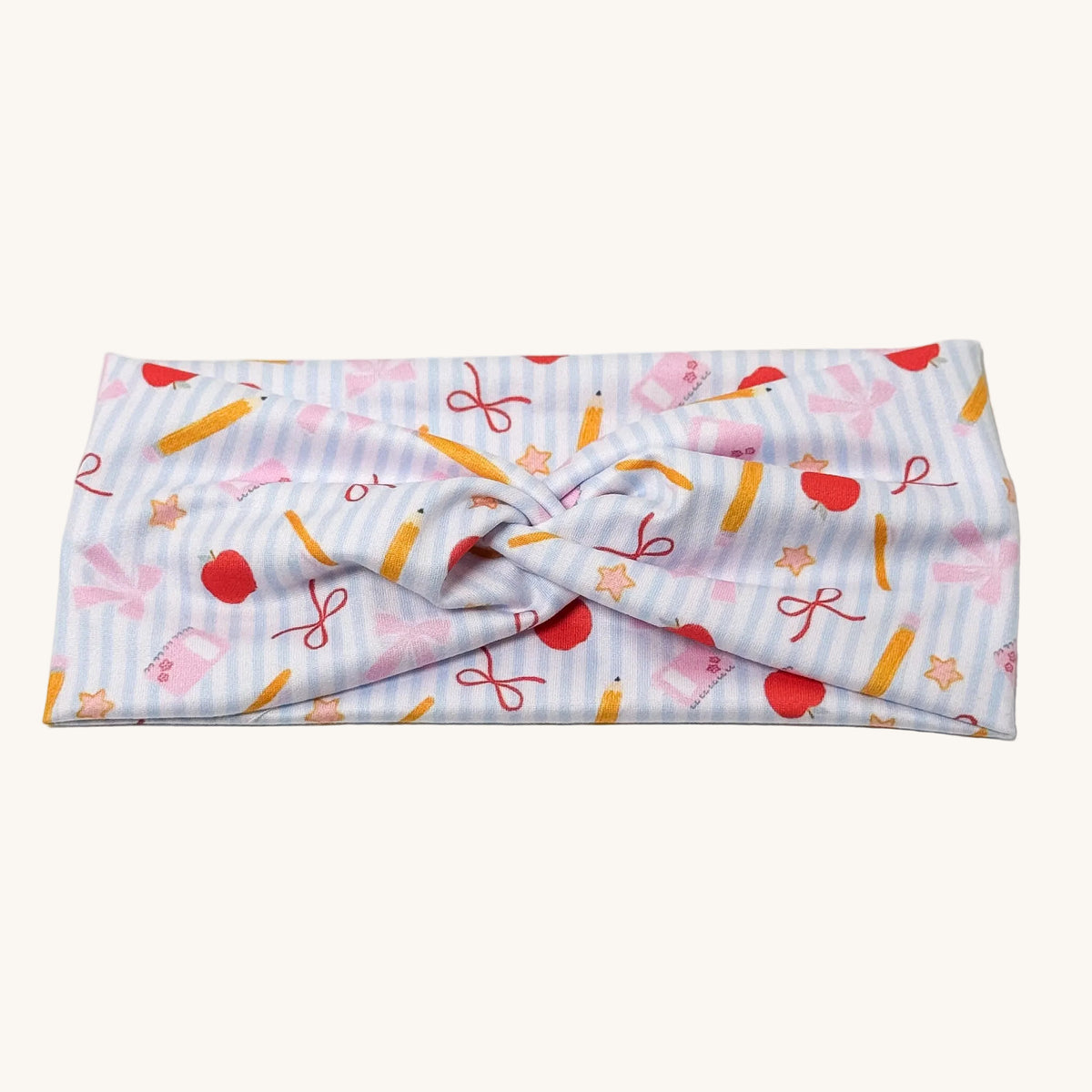 A knotted fabric headband featuring a pastel blue and white striped background with a playful pattern of red apples, yellow pencils, pink bows, and orange stars, displayed on a clean off-white background.
