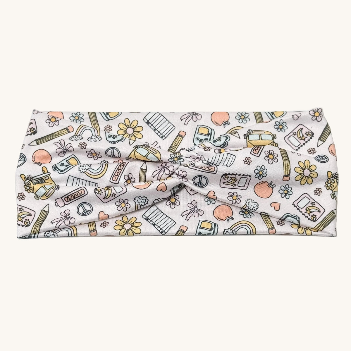 A fabric headband with a twisted center, featuring a colorful doodle-style pattern of school supplies, flowers, and hearts on a white background.