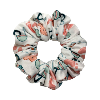 Headband Studio white scrunchie with a baby theme print featuring pink footprints, rattles, and bottles.