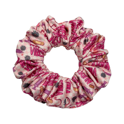 Headband Studio pink scrunchie with nurse vibes print, featuring band-aids, syringes, and flowers. Made of soft polyester.